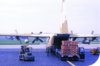 A C-130 transport aircraft on an airfield tarmac with its rear cargo door open. Crates are being loaded onto or offloaded from a truck backed up to the ramp.